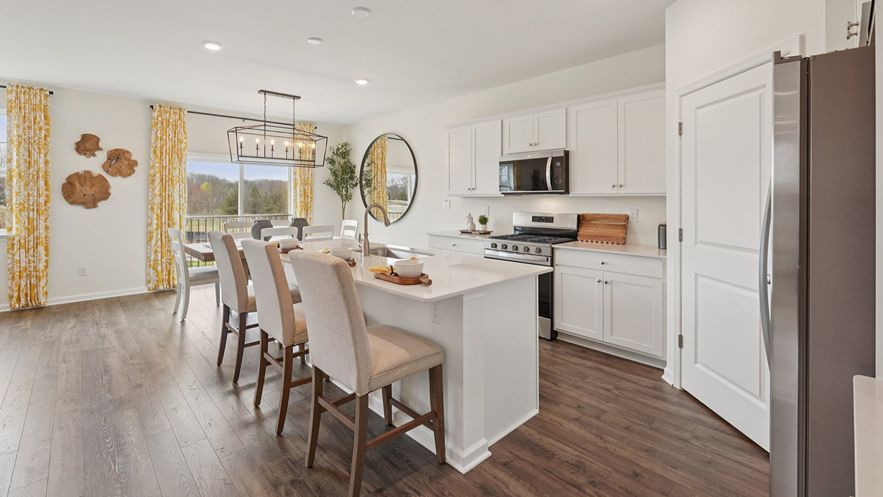 A spacious kitchen with white, shaker-style cabinets, quartz countertops, a large center island with undermount sink and seating for three, and a stainless-steel appliance package overlooking the adjoining dining space and living room.