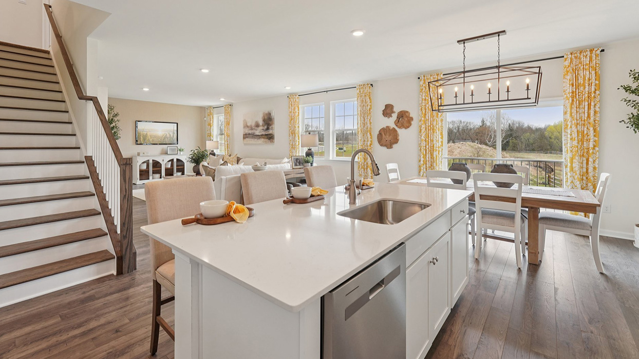 A spacious kitchen with white, shaker-style cabinets, quartz countertops, a large center island with undermount sink and seating for three, and a stainless-steel appliance package overlooking the adjoining dining space and living room.
