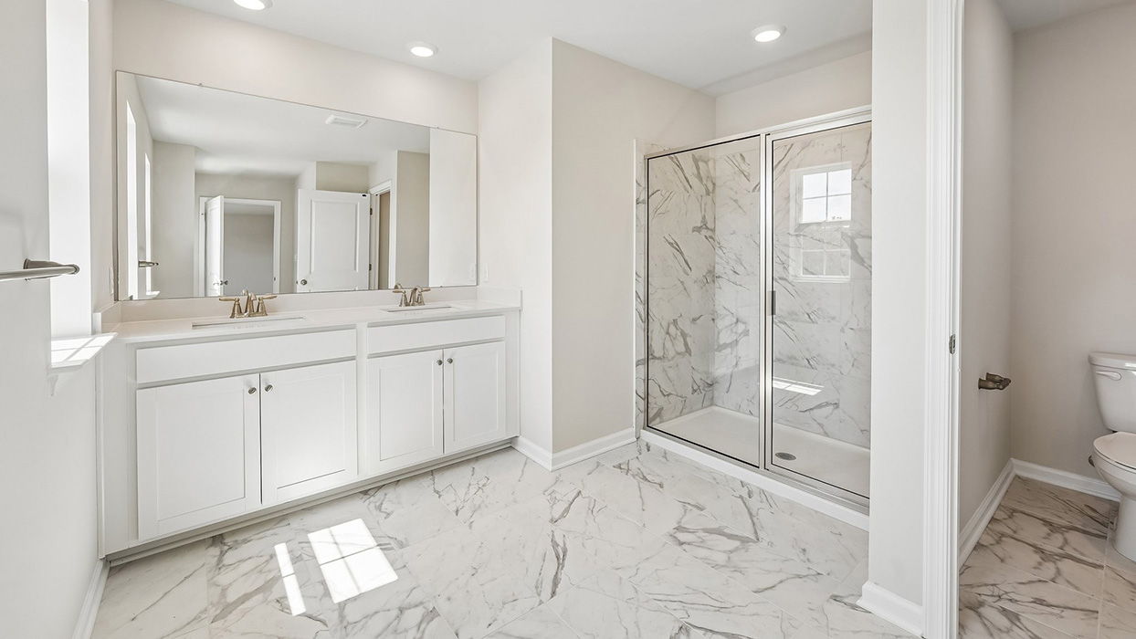 Luxury owner's bathroom in the Galen single-family home at Creekside Village with dual sinks, oversized glass-enclosed shower, private water closet, and marble-look tile flooring.
