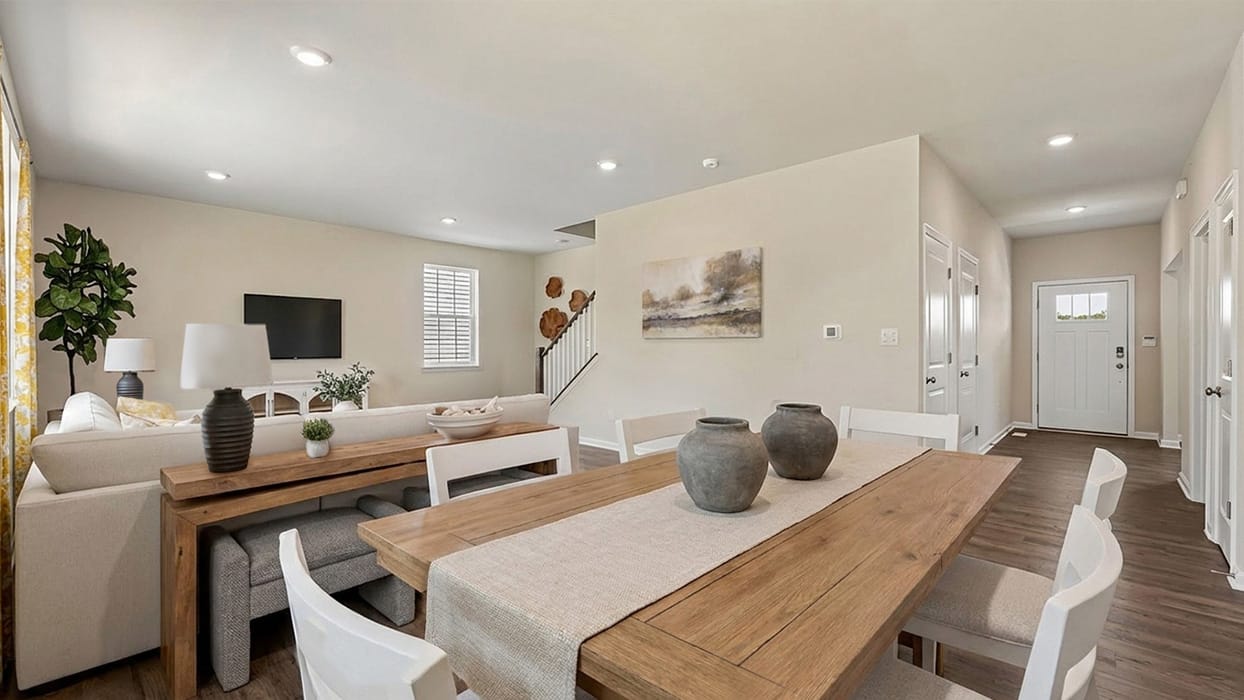 Open-concept dining and living area with a wooden dining table and white chairs in the foreground, a light-colored sectional sofa and wall-mounted TV in the background, neutral walls, recessed lighting, wood-look flooring, and a bright entry hallway leading to the front door.