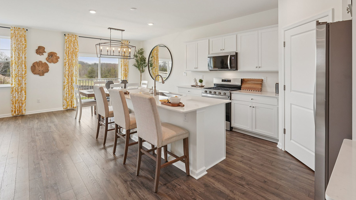 A spacious kitchen with white, shaker-style cabinets, quartz countertops, a large center island with undermount sink and seating for three, and a stainless-steel appliance package overlooking the adjoining dining space and living room.