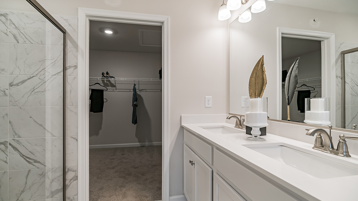 Connecting primary bath with double sink white vanity and white and gray marble tile shower with connecting closet.