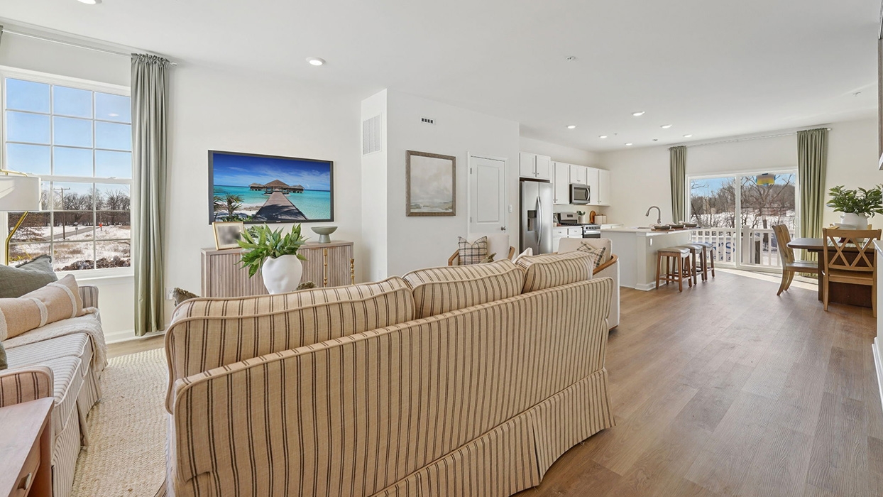 Open-concept living room looking into kitchen and dining area, featuring a large sofa and sliding glass door for natural light