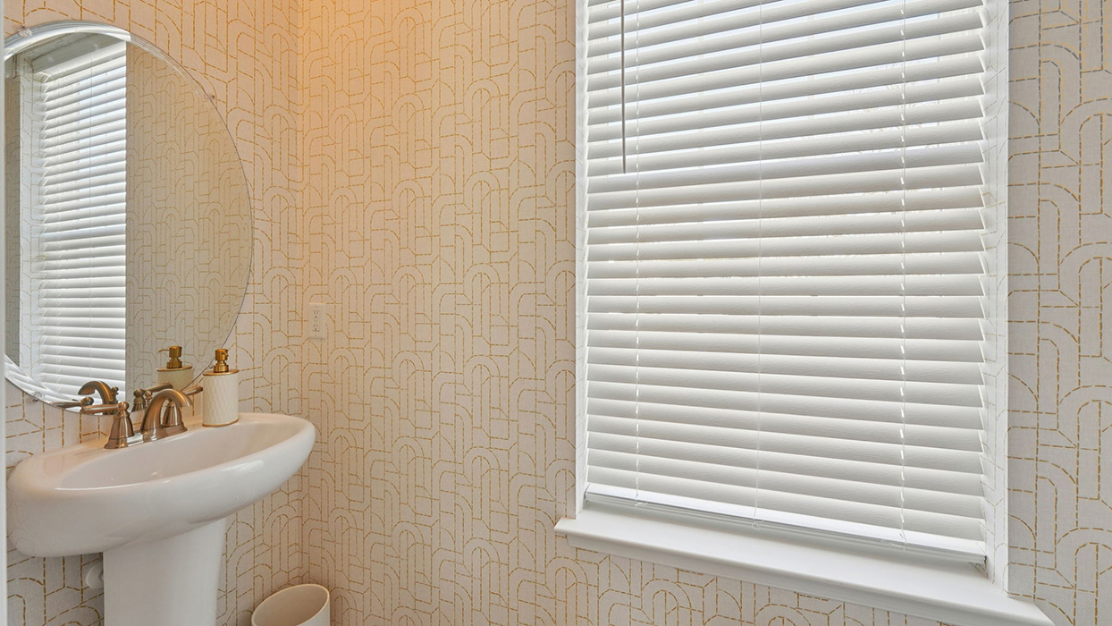 Powder room with pedestal sink, round mirror, and patterned wallpaper with natural light from window