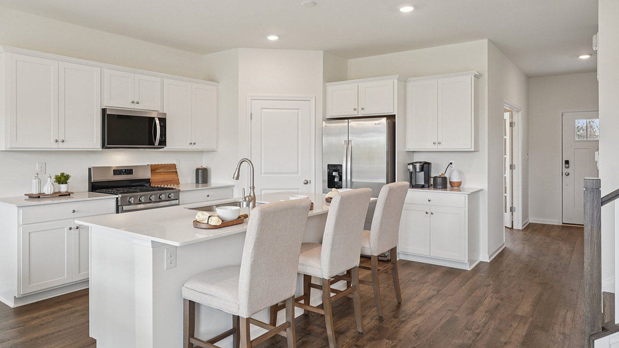 A spacious kitchen with white, shaker-style cabinets, quartz countertops, a large center island with undermount sink and seating for three, and a stainless-steel appliance package overlooking the adjoining dining space and living room.