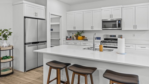 Another view of the kitchen at the Stillwater model home in Long Valley with white cabinets and white expansive countertops, stainless steel appliances and a large pantry.