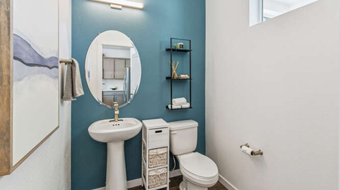 Modern powder room of furnished Muirfield model home featuring a pedestal sink, oval mirror, LED light fixture and clerestory window