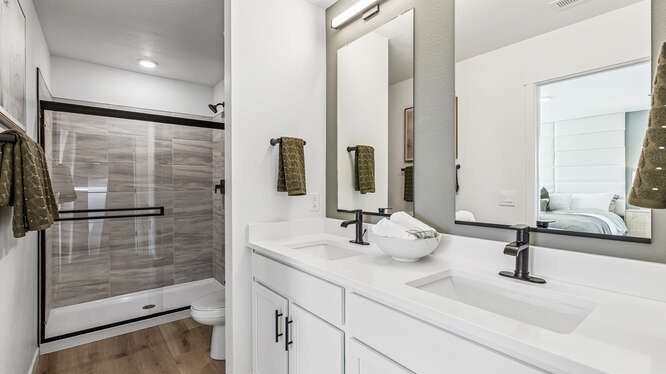 Furnished Muirfield model home private bathroom with dual sink vanity and white cabinetry in foreground and large glass-enclosed shower