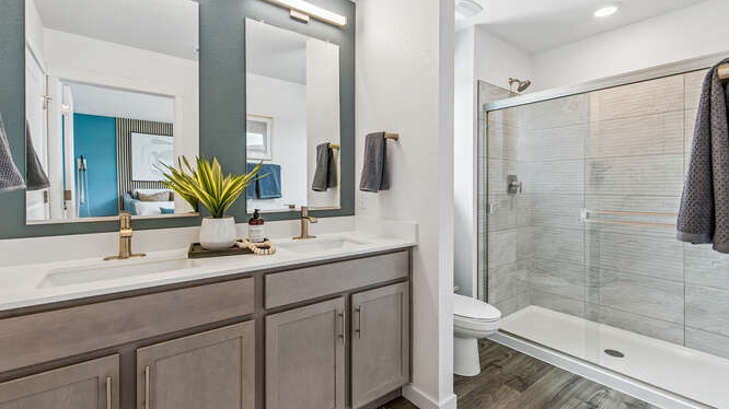 Furnished Muirfield model home primary private bathroom with dual sink vanity and cabinetry in foreground and large glass-enclosed shower