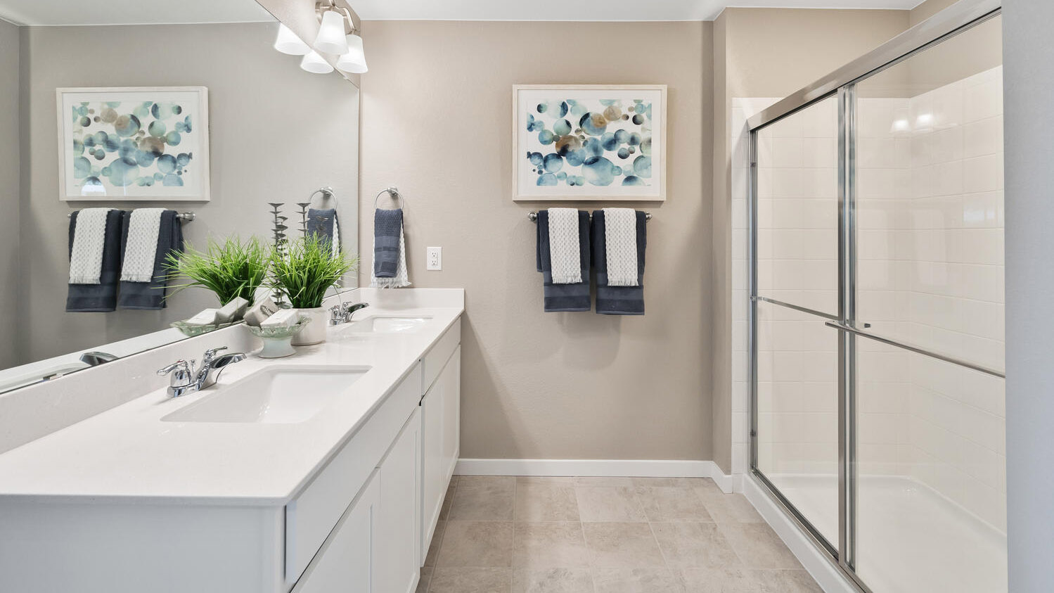 Galley view of private bathroom in primary bedroom suite showing step-in shower and double sinks with vanity lighting