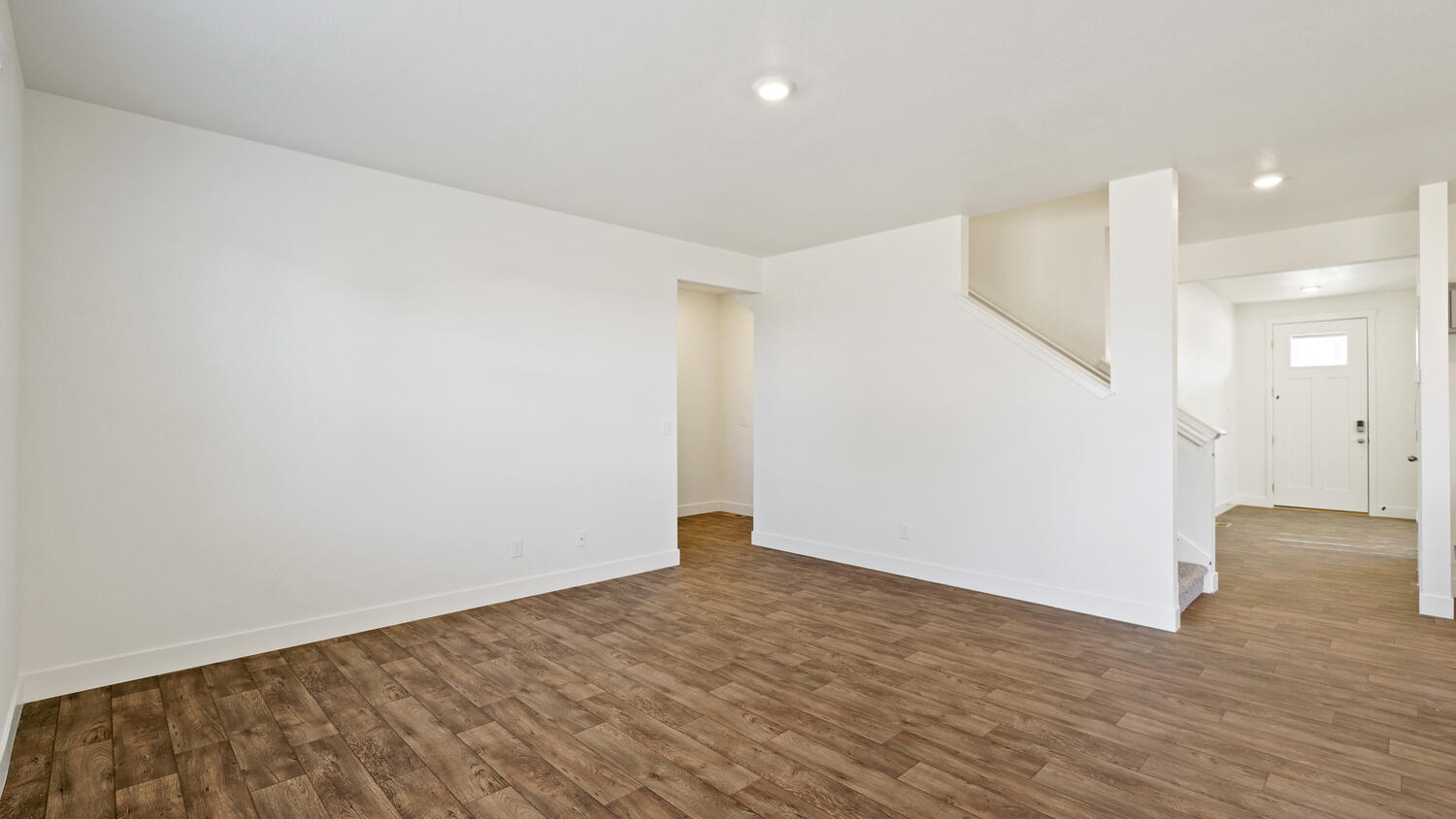 Alternate angle of great room showing hallway entrance to full bathroom and secondary bedroom, stairwell leading to upper floor, and front door