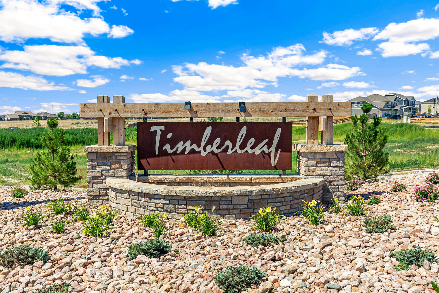 Artful rustic and industrial mixed-media Timberleaf community monumet sign on a plant-laden bed of stones, new homes in background.