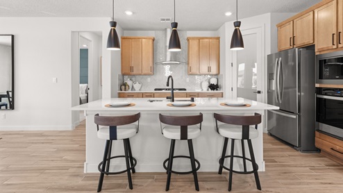 The Destin Kitchen with brown tile floors, white backsplash, kitchen island with granite countertops, and three pendant lights over the island.