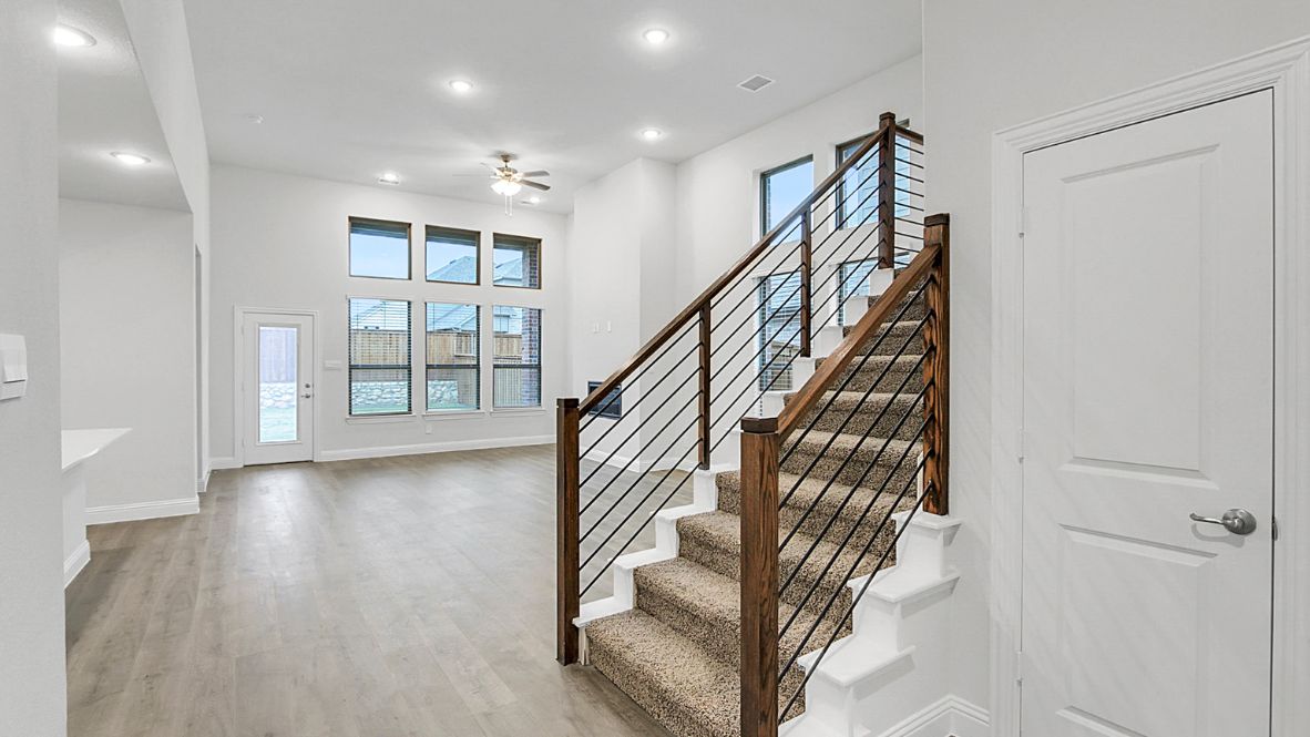 Modern open-concept living room with cozy fireplace, large windows offering natural light, and wood-look flooring. The space features a contemporary staircase with sleep railing leading to an upper loft area, and a view of the bright kitchen with white cabinetry, stainless steel appliances, and a spacious island.