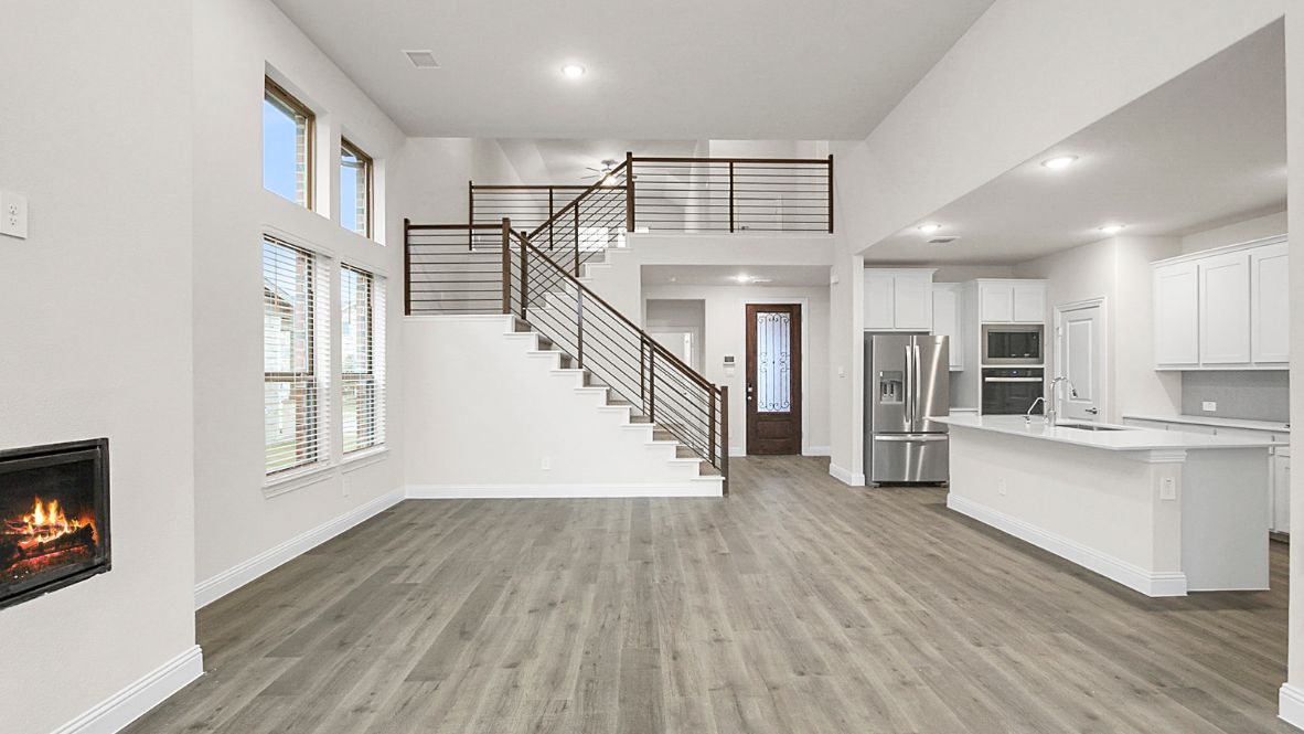 Modern open-concept living room with cozy fireplace, large windows offering natural light, and wood-look flooring. The space features a contemporary staircase with sleep railing leading to an upper loft area, and a view of the bright kitchen with white cabinetry, stainless steel appliances, and a spacious island.