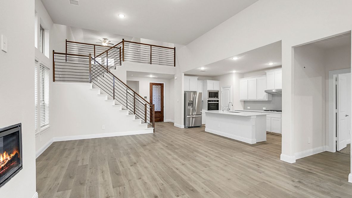 Modern open-concept living room with cozy fireplace, large windows offering natural light, and wood-look flooring. The space features a contemporary staircase with sleep railing leading to an upper loft area, and a view of the bright kitchen with white cabinetry, stainless steel appliances, and a spacious island.