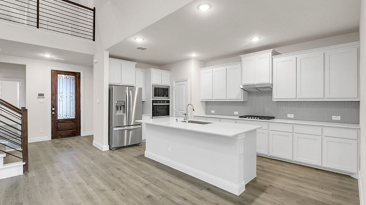 Bright and spacious open-concept kitchen and living area featuring modern white cabinetry, a large center island, and wood-look flooring. The design includes a loft with sleek railing, a high ceiling with ceiling fans, and carpeted stairs leading to the upper level.