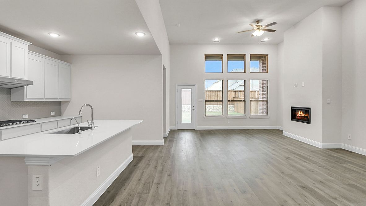 Open-concept kitchen and living area featuring a modern island with a stainless-steel sink and dishwasher, white cabinetry, and built-in appliances. The living room boasts a cozy fireplace, large windows allowing natural light, and wood-look flooring, creating a warm and inviting atmosphere.