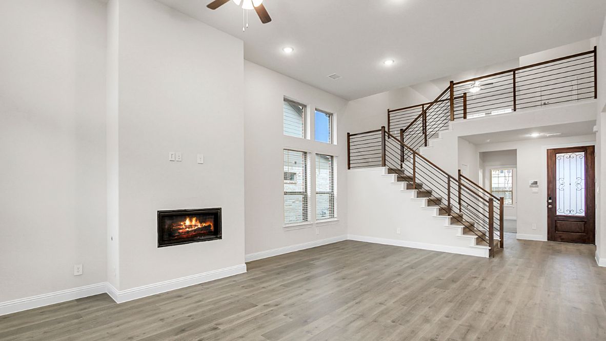 Modern open-concept living room with cozy fireplace, large windows offering natural light, and wood-look flooring. The space features a contemporary staircase with sleep railing leading to an upper loft area.