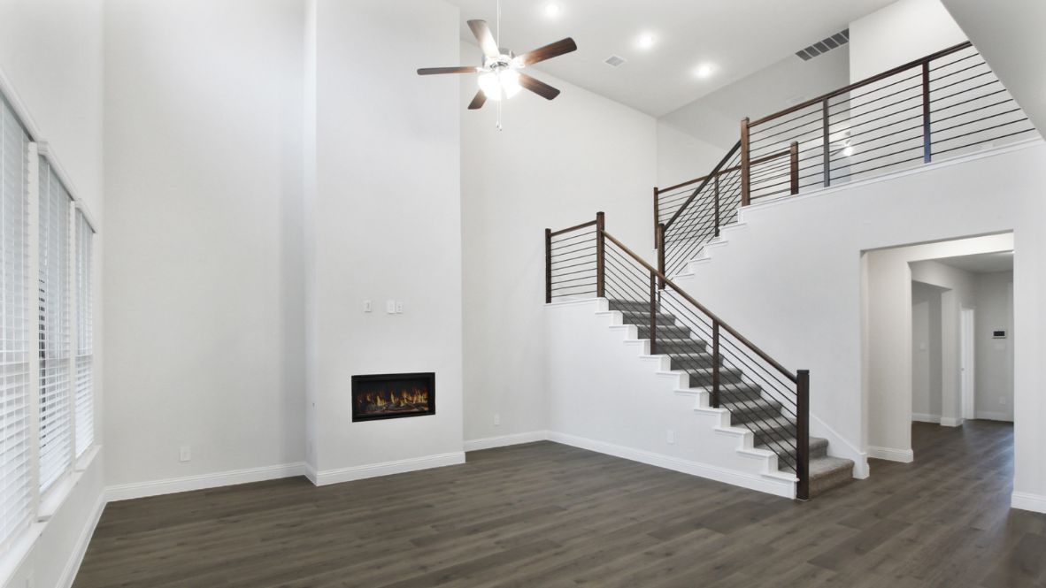 Modern open-concept living room with cozy fireplace, large windows offering natural light, and wood-look flooring. The space features a contemporary staircase with sleep railing leading to an upper loft area, and a view of the bright kitchen with white cabinetry, stainless steel appliances, and a spacious island.