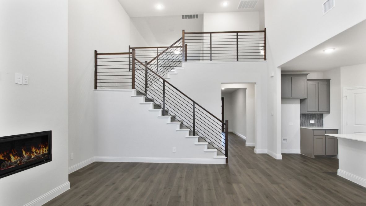 Modern open-concept living room with cozy fireplace, large windows offering natural light, and wood-look flooring. The space features a contemporary staircase with sleep railing leading to an upper loft area, and a view of the bright kitchen with white cabinetry, stainless steel appliances, and a spacious island.